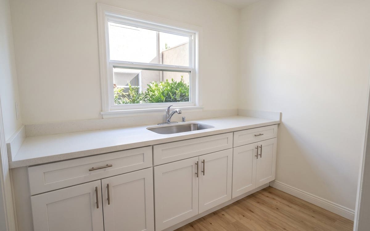 Apartment cleaning tips start in a small kitchenette: clear white counter, stainless sink and a window onto courtyard greenery.