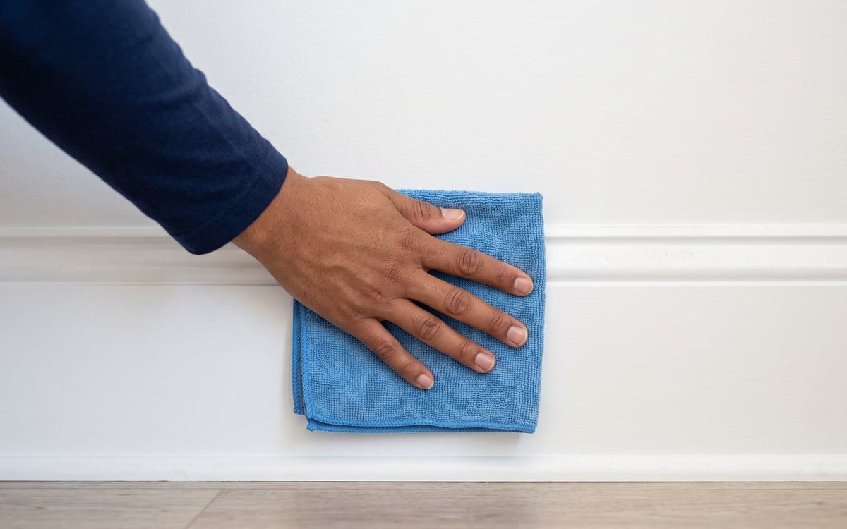Along a white baseboard, a bare hand presses a folded blue microfiber cloth during a deep cleaning service.