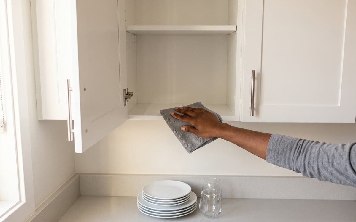 Plates and glasses stacked on the counter below, a bare hand wipes the shelf of an emptied upper cabinet during a deep cleaning service.