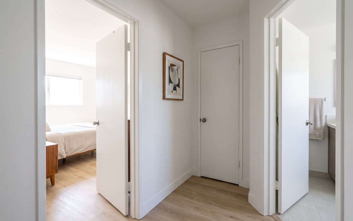 House cleaning cost for a one-bedroom: a short hallway with the bedroom and bathroom doors open, light plank floors and a framed print.