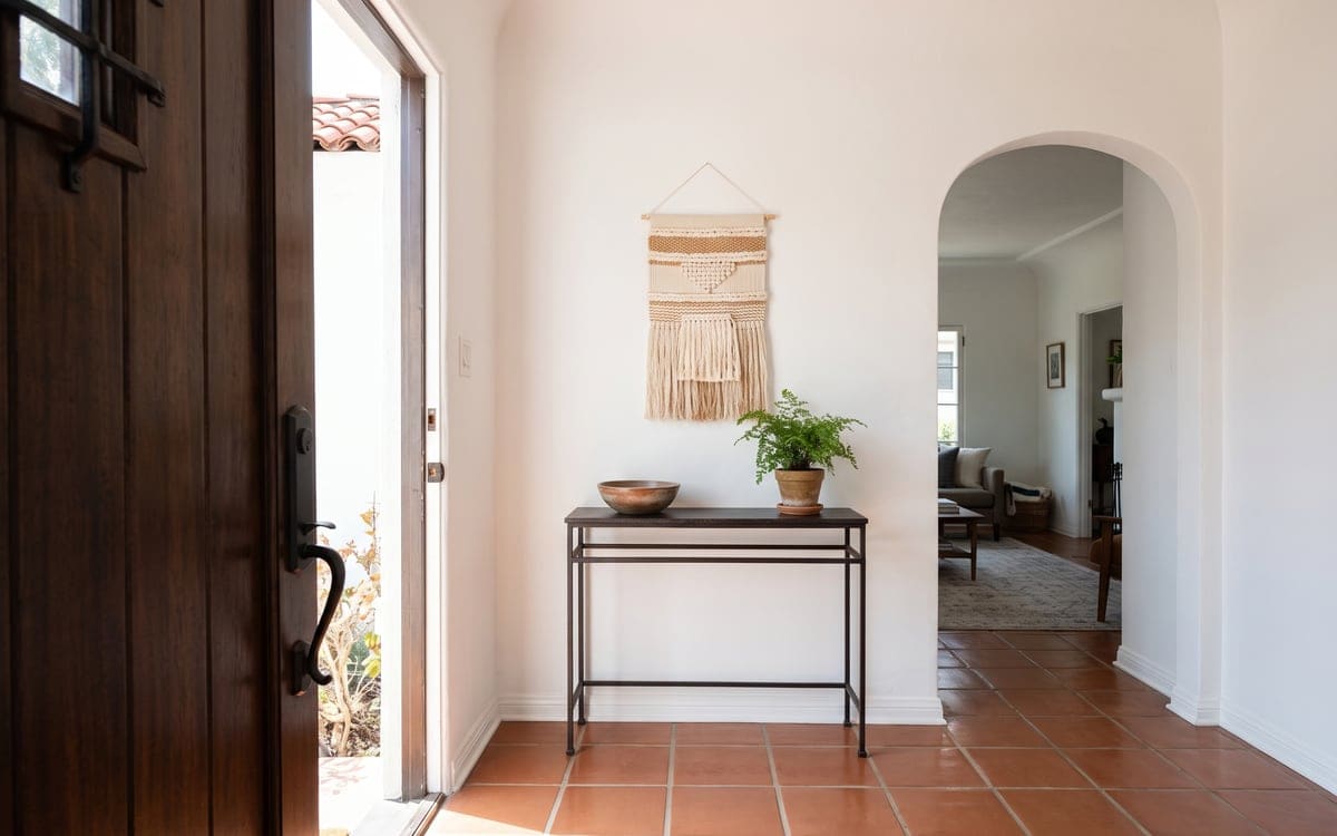 A Spanish revival entry hall after cleaning services, with a terracotta tile floor, an arched doorway, a dark console with a fern and the wood front door open.