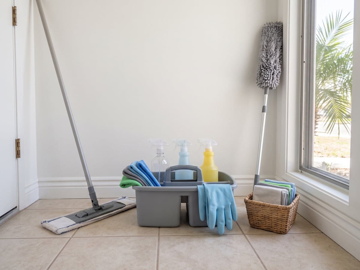 A caddy of spray bottles, flat mop, duster and gloves on a bright tile entry floor, the kit for cleaning services at a Los Angeles home.